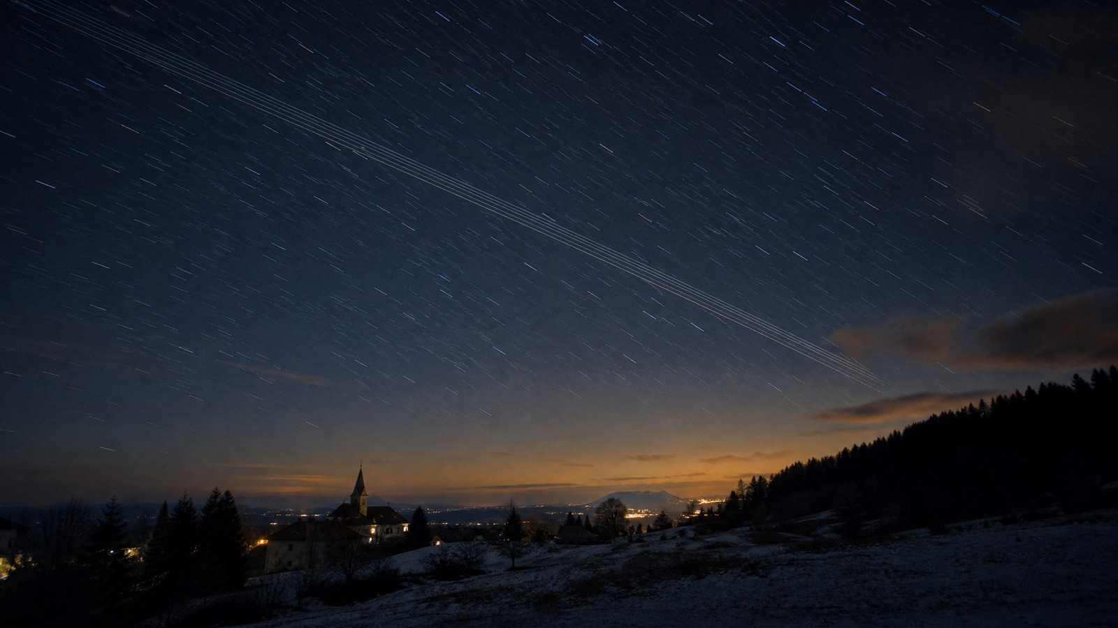 Long-exposure night sky over a European alpine village showing star trails and parallel satellite streaks