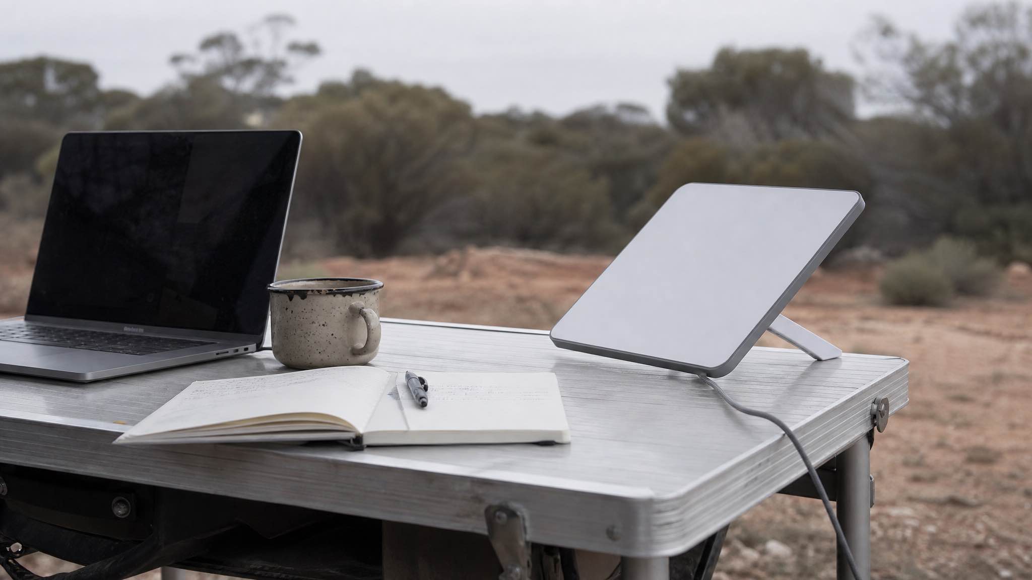 Laptop on a fold-out camping table next to a portable satellite dish on red dirt in the Western Australian outback at golden hour
