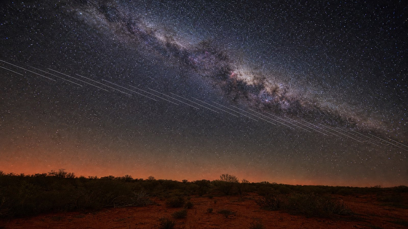 Long-exposure photo of the Milky Way over remote Australian outback scrub, crossed by parallel white satellite trails from a Starlink train.