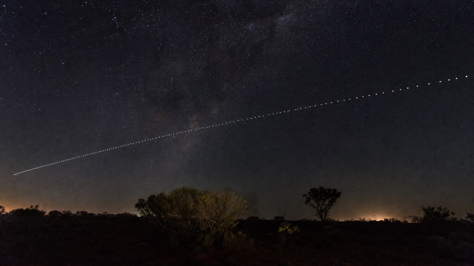 Long-exposure night sky photograph showing a Starlink satellite train crossing the stars above remote bushland