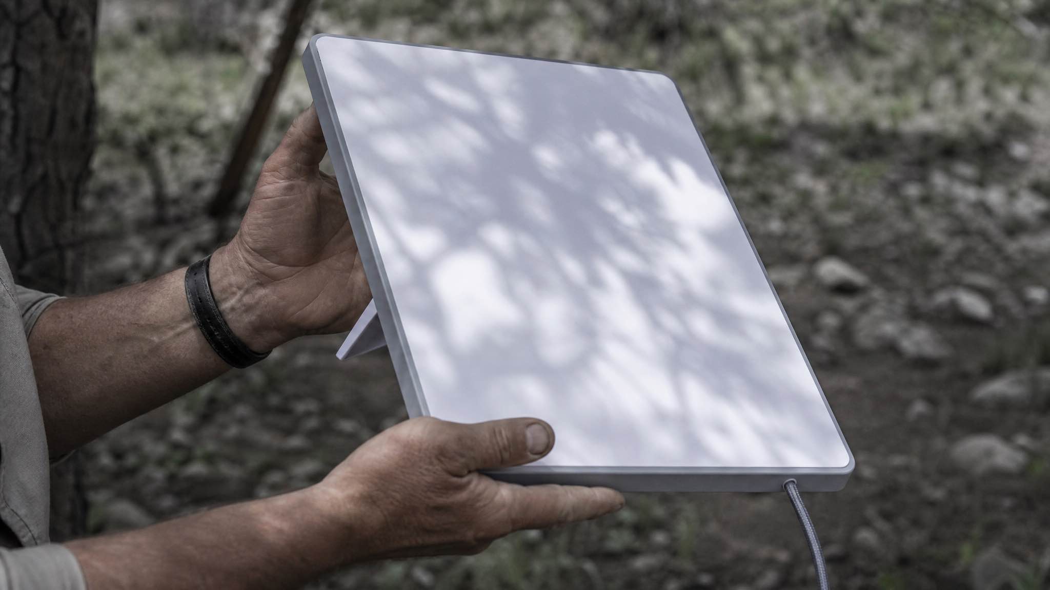 Hands inspecting the back of a Starlink Mini dish on a camp table in the WA bush, tree shadow falling across the panel